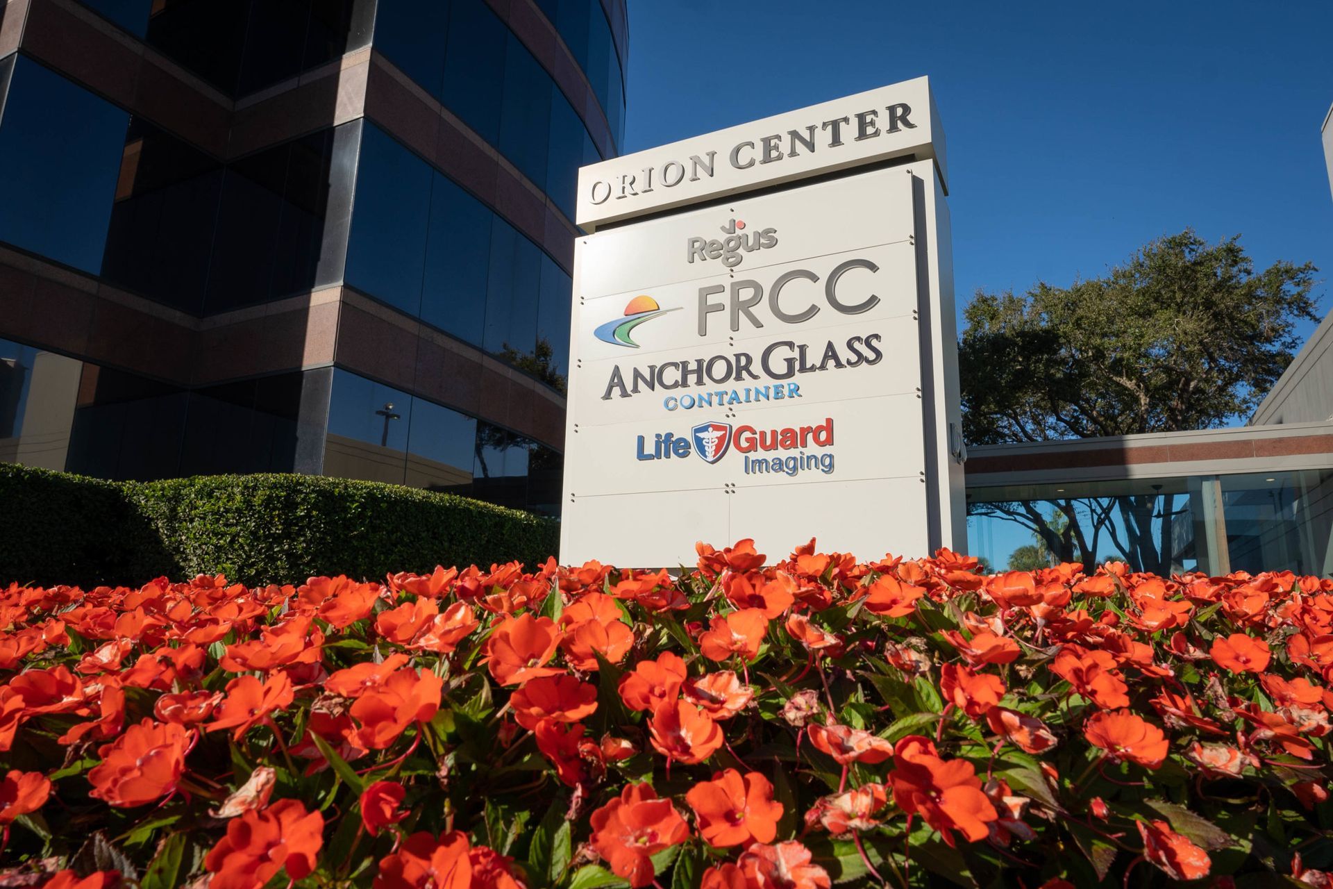 Sign for Orion Center with company names: FRCC, Anchor Glass, and UltraGuard Imaging, with red flowers. Sign for Orion Center with company names: FRCC, Anchor Glass, and UltraGuard Imaging, with red flowers.