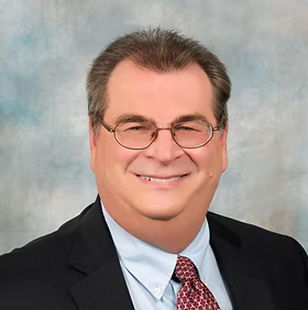 Man in glasses, suit, and tie smiling, against a blurred blue and white background.
