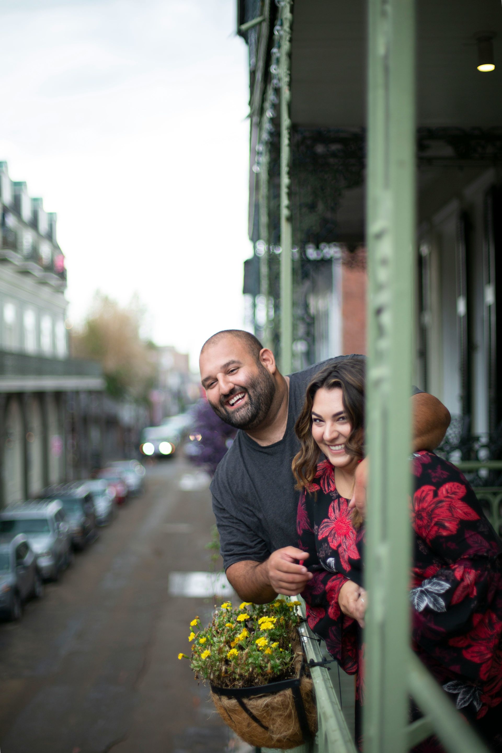 Couple smiling on a balcony overlooking a city street. Balcony has hanging flowers.