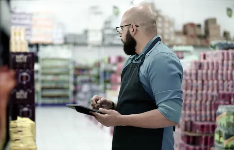 Man in apron using a tablet in a supermarket, checking shelves with packaged goods.