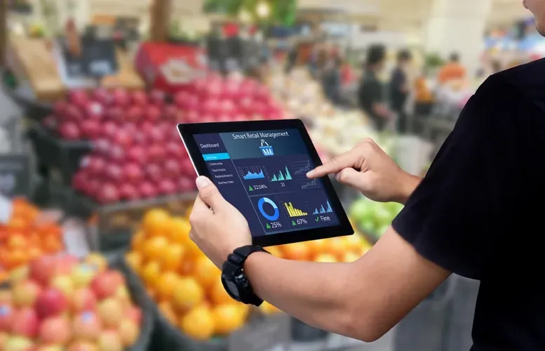 Person using a tablet in a grocery store, displaying data charts, fruits in the background.