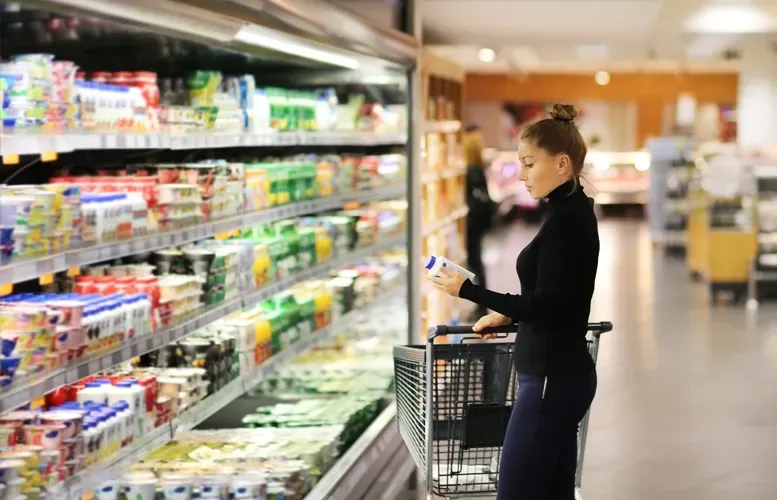 Woman shopping for dairy products in supermarket, holding a yogurt container next to shopping cart.