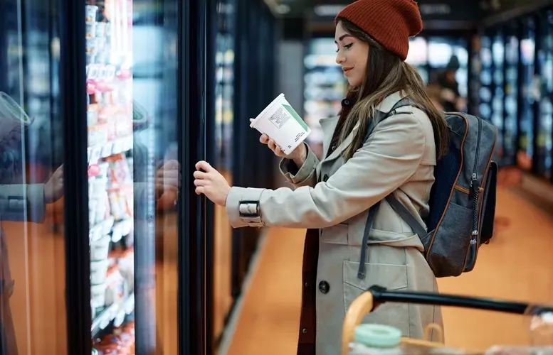 A woman in a coat and hat at a grocery store freezer, holding a product and looking inside.