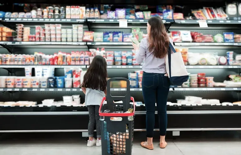 A woman and a child are shopping in a grocery store, looking at refrigerated items. A shopping cart sits in front of them.