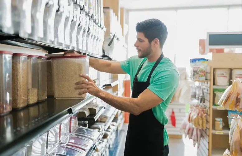 Man in black apron restocking shelf in a store, holding a container of grains.