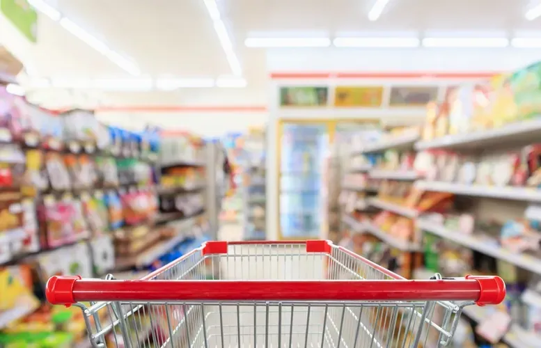 Red-handled shopping cart in a grocery store aisle, shelves stocked with products, blurry background.