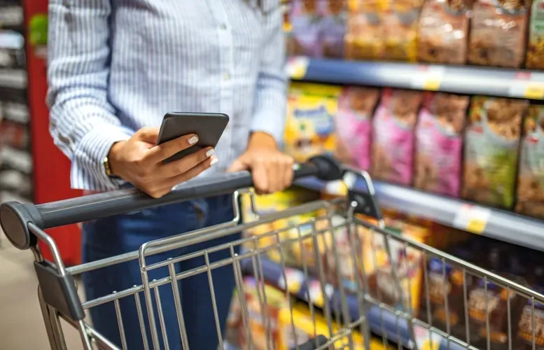 A person with a smartphone and a shopping cart in a grocery store aisle, looking at product shelves.