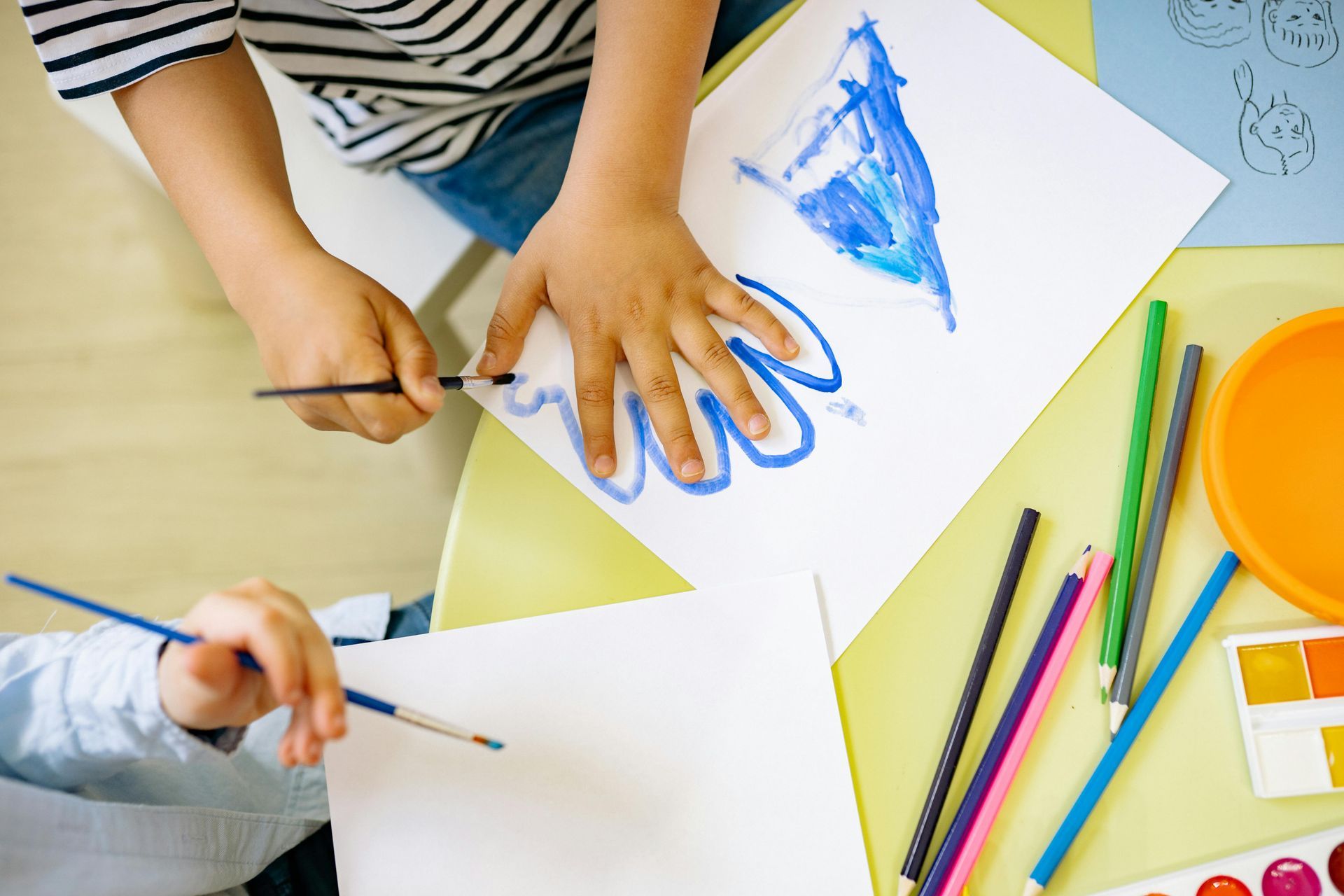 Child using paintbrushes to color on white paper at a table, with crayons and paint set.