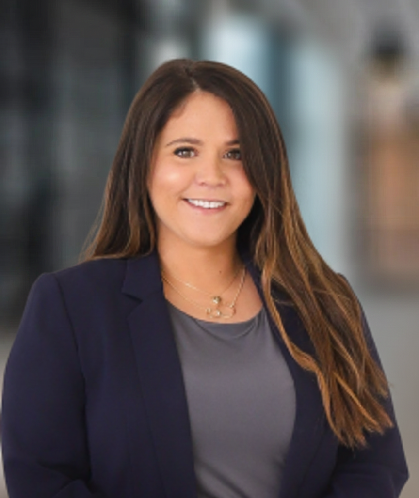Woman with long brown hair smiles, wearing a navy blazer and gray top, blurred background.