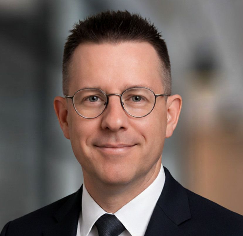Man with glasses wearing a dark suit and tie, smiling, in a blurred office setting.