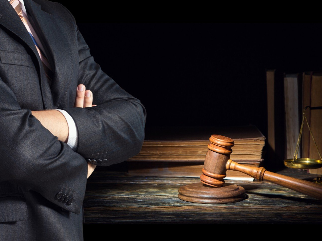 Uomo in giacca e cravatta con le braccia incrociate, accanto a martelletto, libri e bilancia su una scrivania di legno. Sfondo scuro.