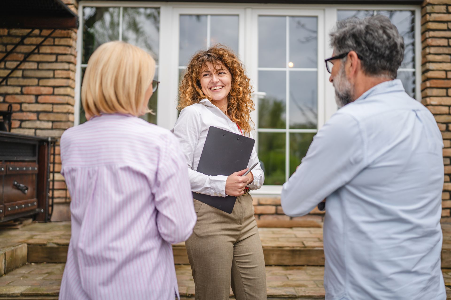 Real estate agent smiles while speaking with a couple in front of a brick home.