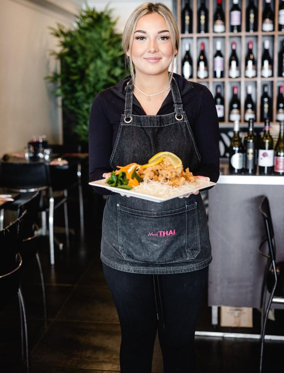 A Woman In An Apron Is Holding A Plate Of Food In A Restaurant — Mod Thai Nelson Bay In Nelson Bay, NSW