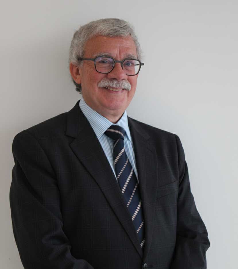 Man in suit and tie, smiling, wearing glasses, against a plain white wall.