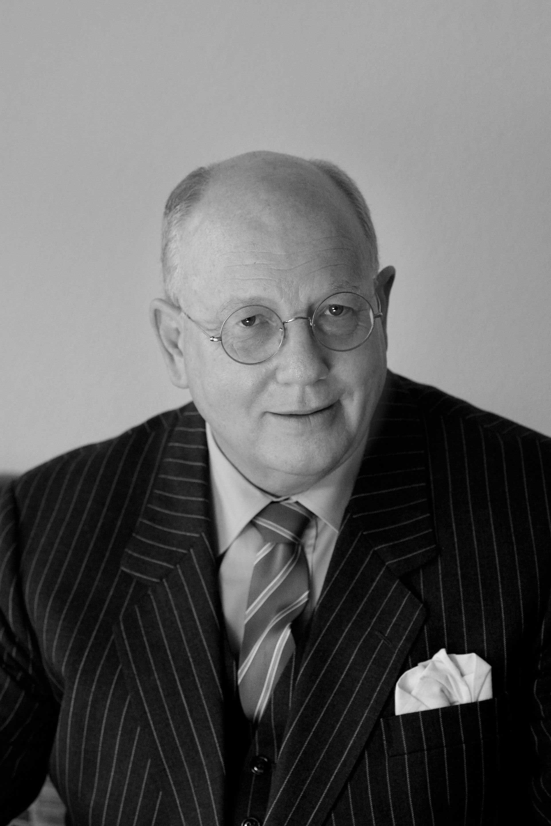 A man in a pinstripe suit and glasses, looking at the camera. He wears a tie and has a handkerchief in his pocket.