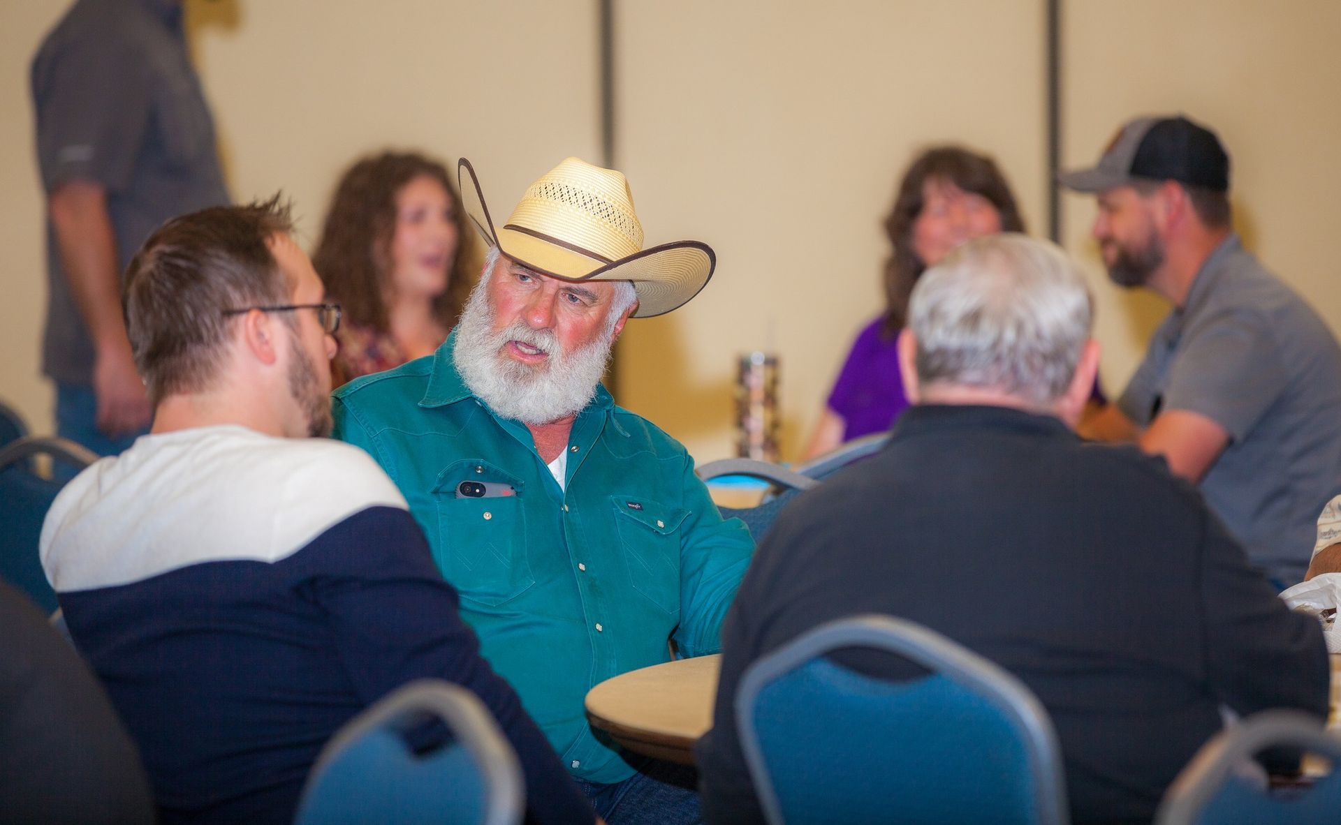 A group of people are sitting around a table talking to each other . one of the men is wearing a cowboy hat.
