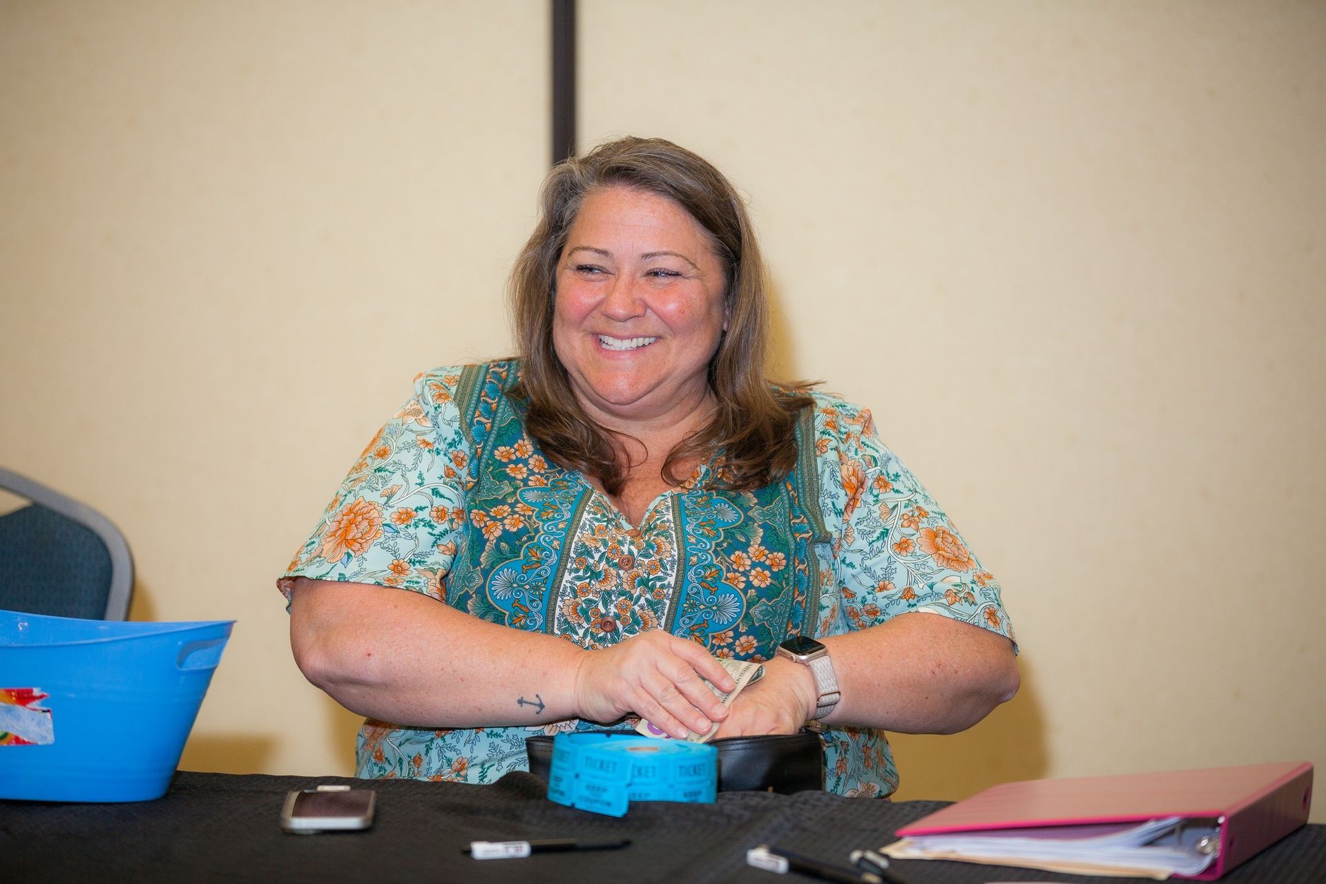 A woman is sitting at a table with her hands folded and smiling.