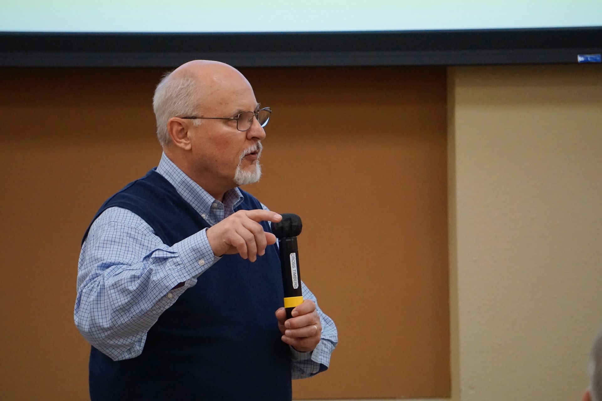 A man with glasses and a beard is holding a microphone and pointing at it.