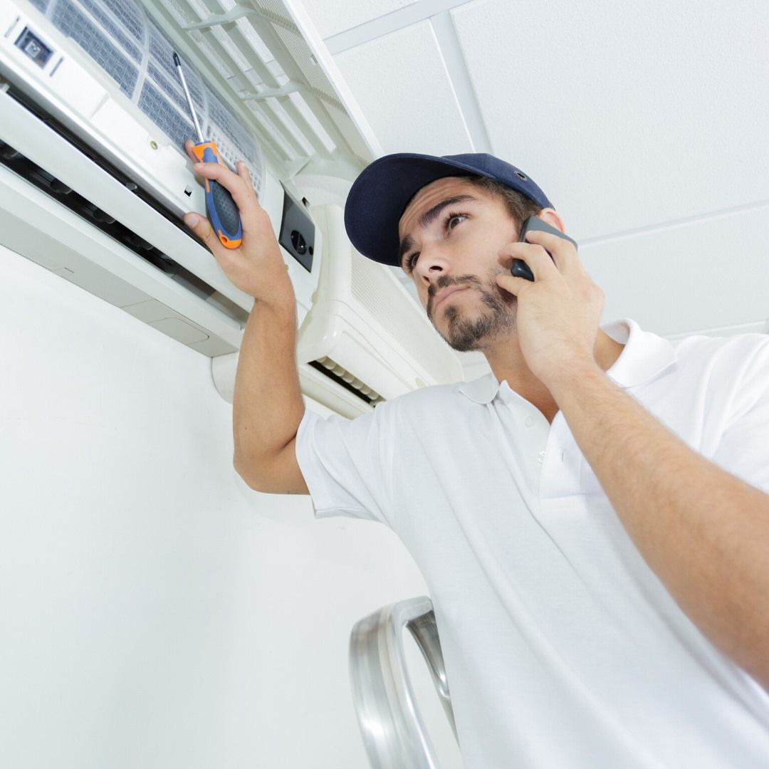 A man is fixing an air conditioner while talking on a cell phone