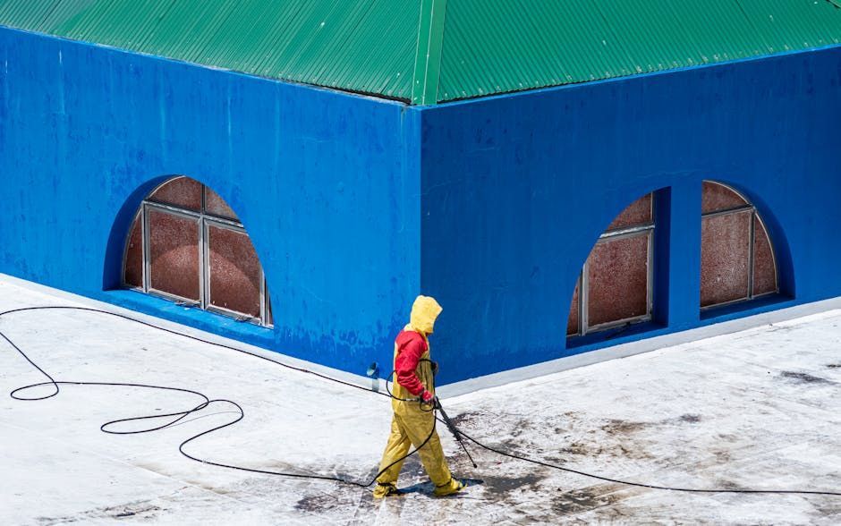 Person in yellow safety gear power-washing a white roof, blue building with arched windows, and a green roof visible. Person in yellow suit power washes a white roof with blue and green building.