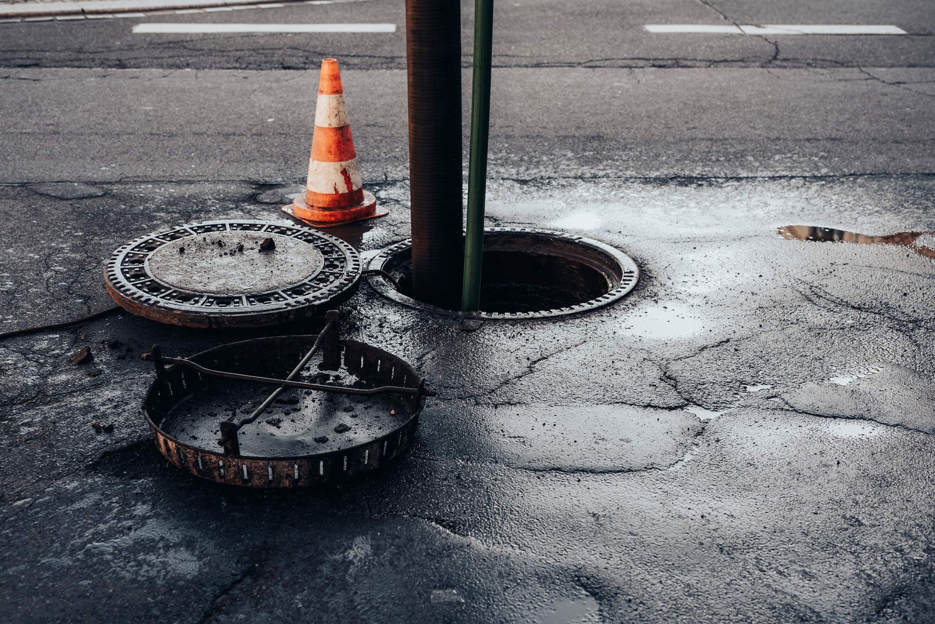 Open manhole in cracked pavement with cone, cover, and pole. Gray asphalt, wet.