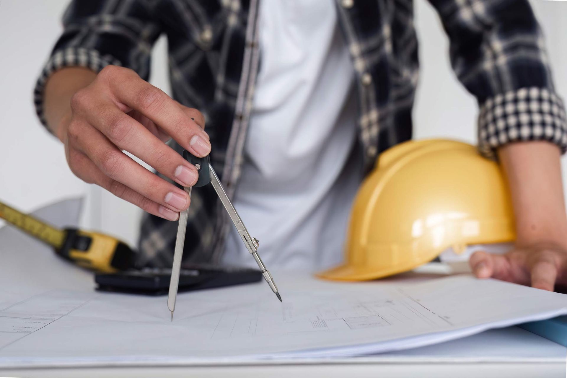 Person using a compass on a blueprint, next to a hard hat and calculator.