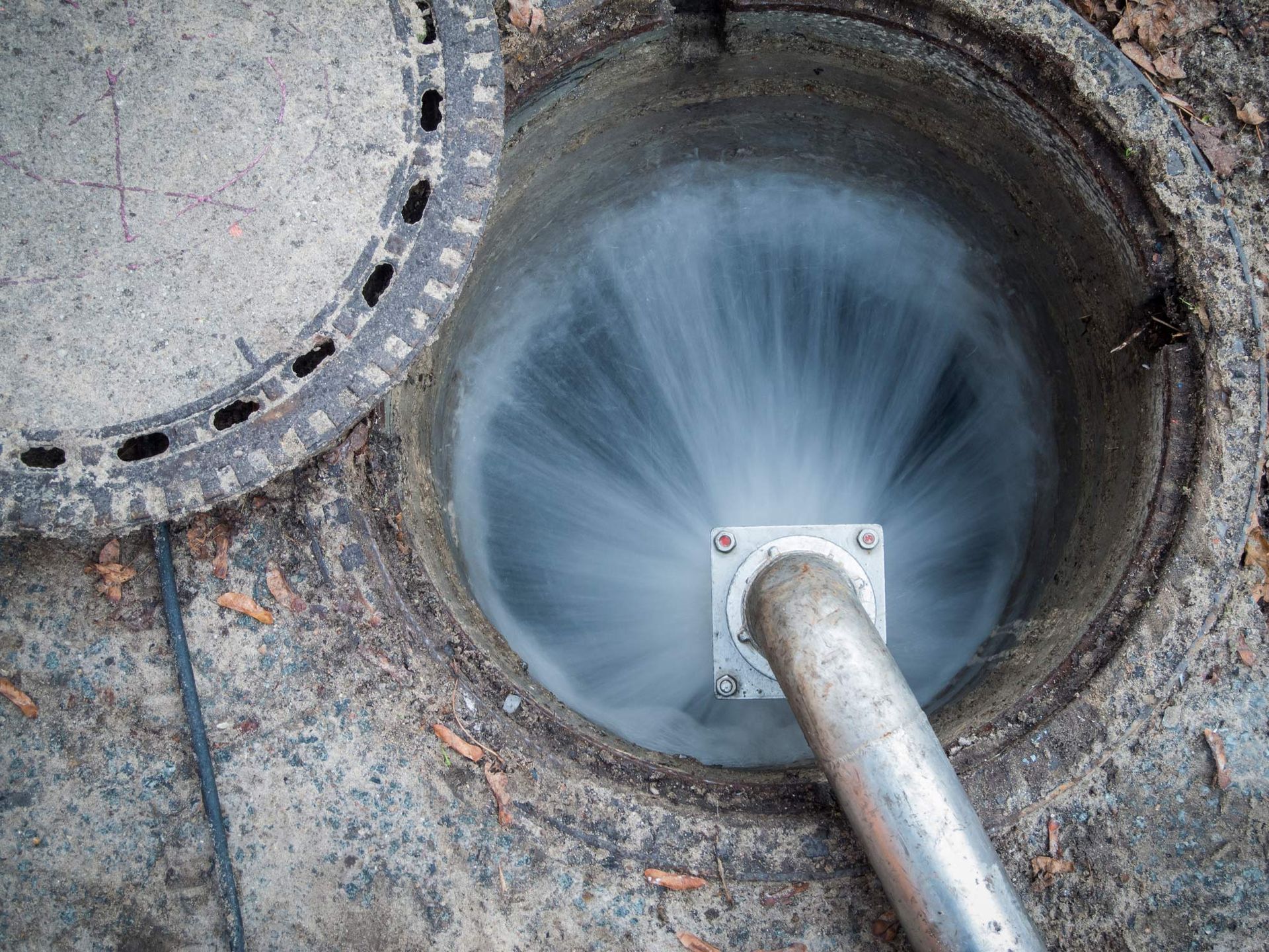 Water gushing forcefully from a silver pipe into an open manhole, with the lid partially open.