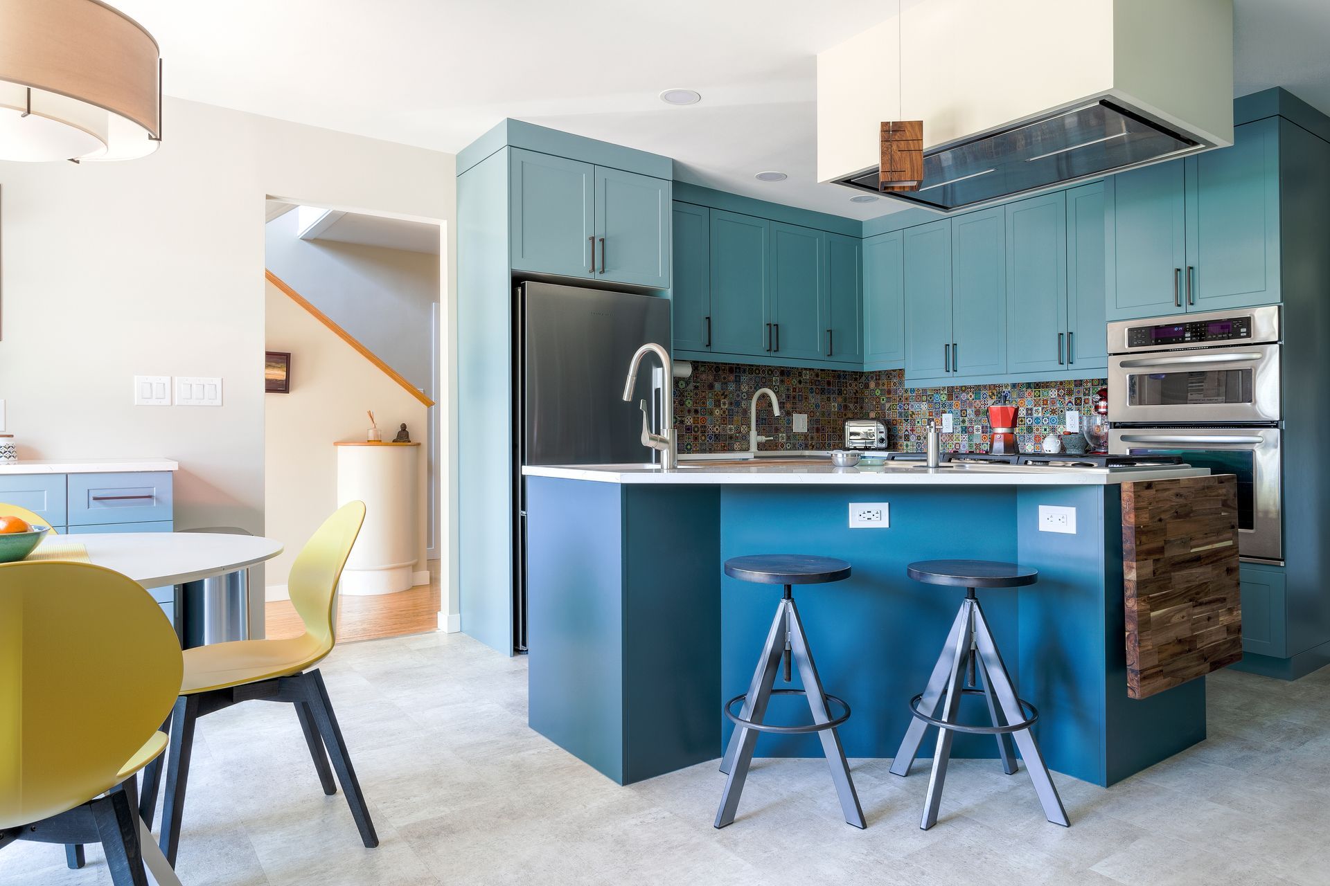 A kitchen with blue cabinets , stools , a table and a refrigerator.