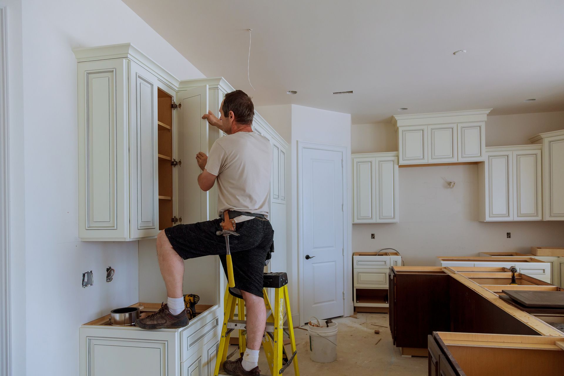 A man is standing on a ladder fixing cabinets in a kitchen.