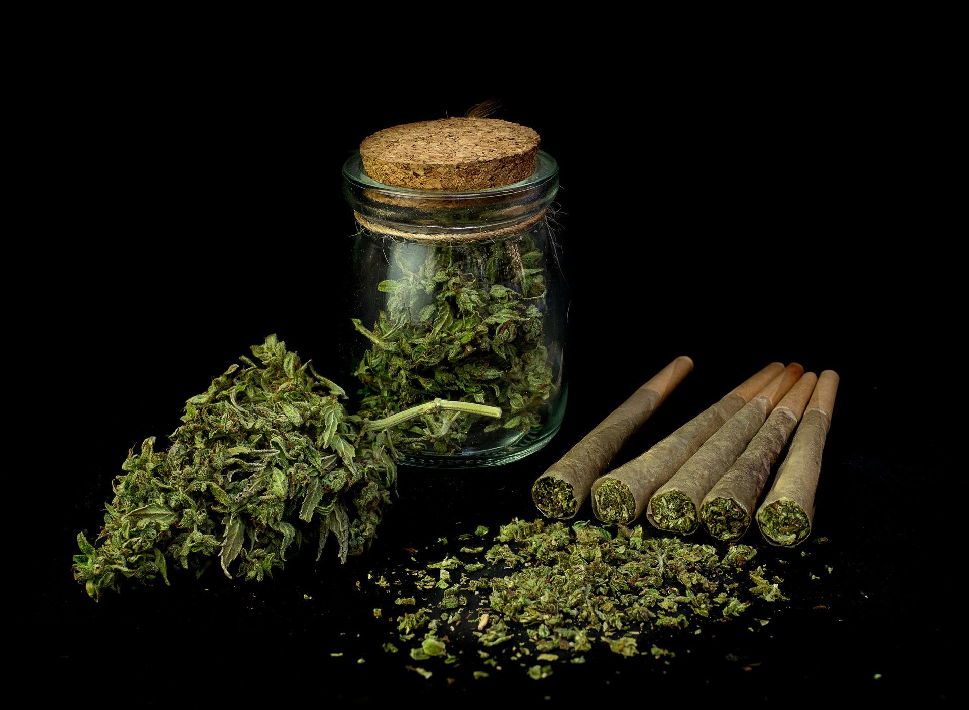 Glass jar filled with cannabis buds next to dried cannabis flower and five pre-rolled joints on a black background.