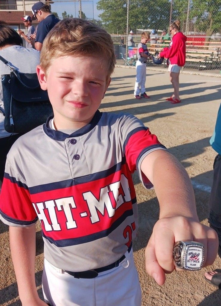 A young boy wearing a hit-man jersey is holding a baseball