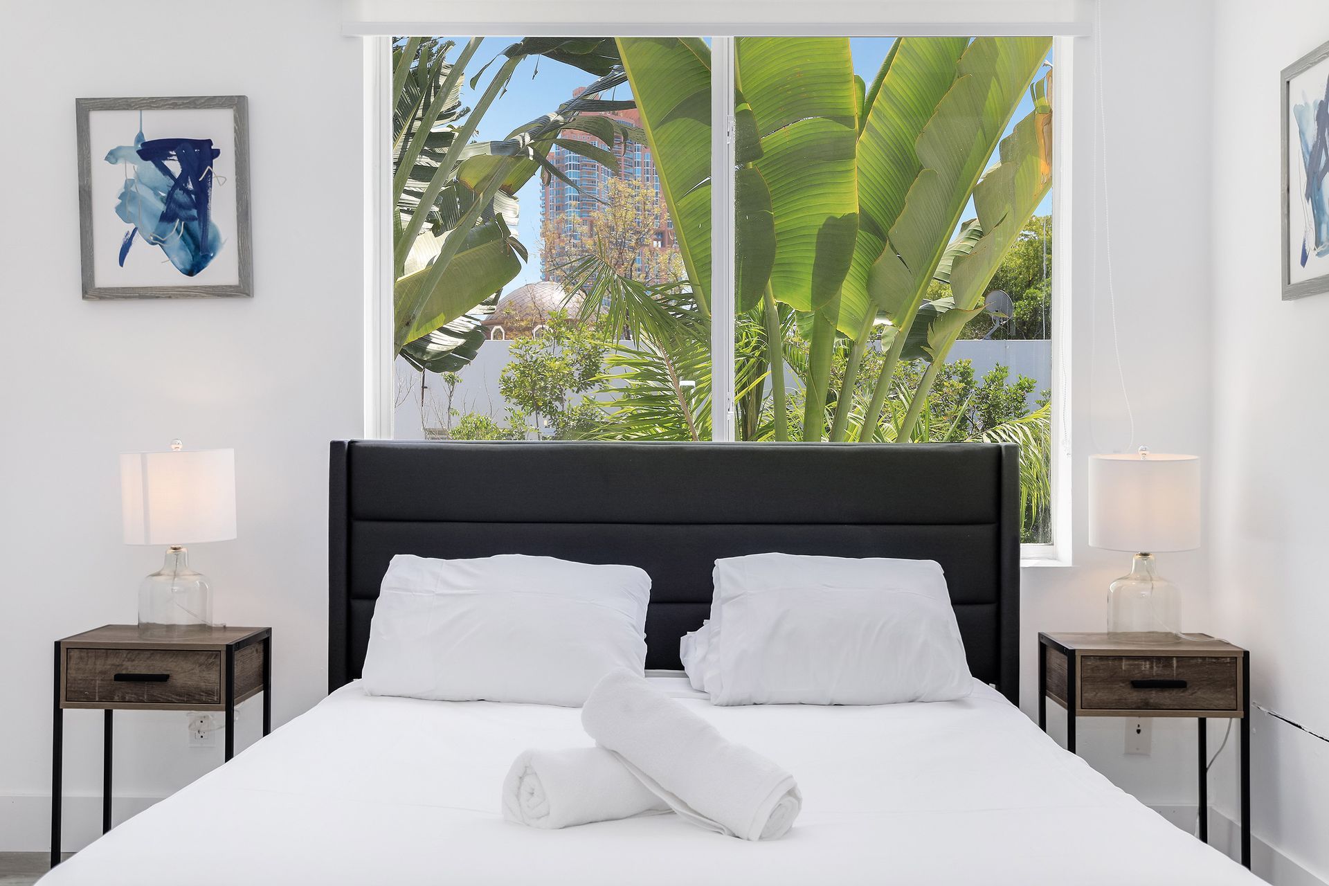 Bedroom with a bed, nightstands, and a window overlooking greenery. White walls and artwork.