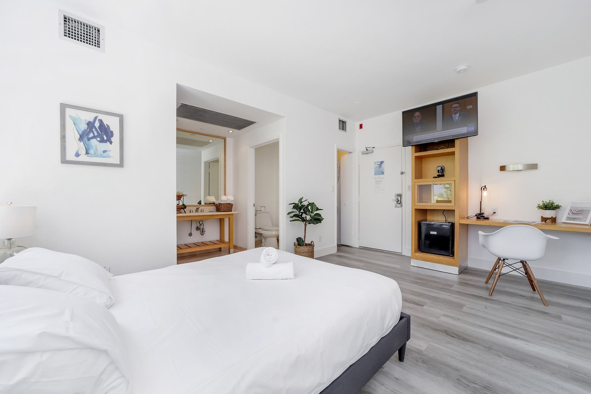 Modern hotel room with a bed, desk, and bookshelf. White walls, gray floors, and natural light.