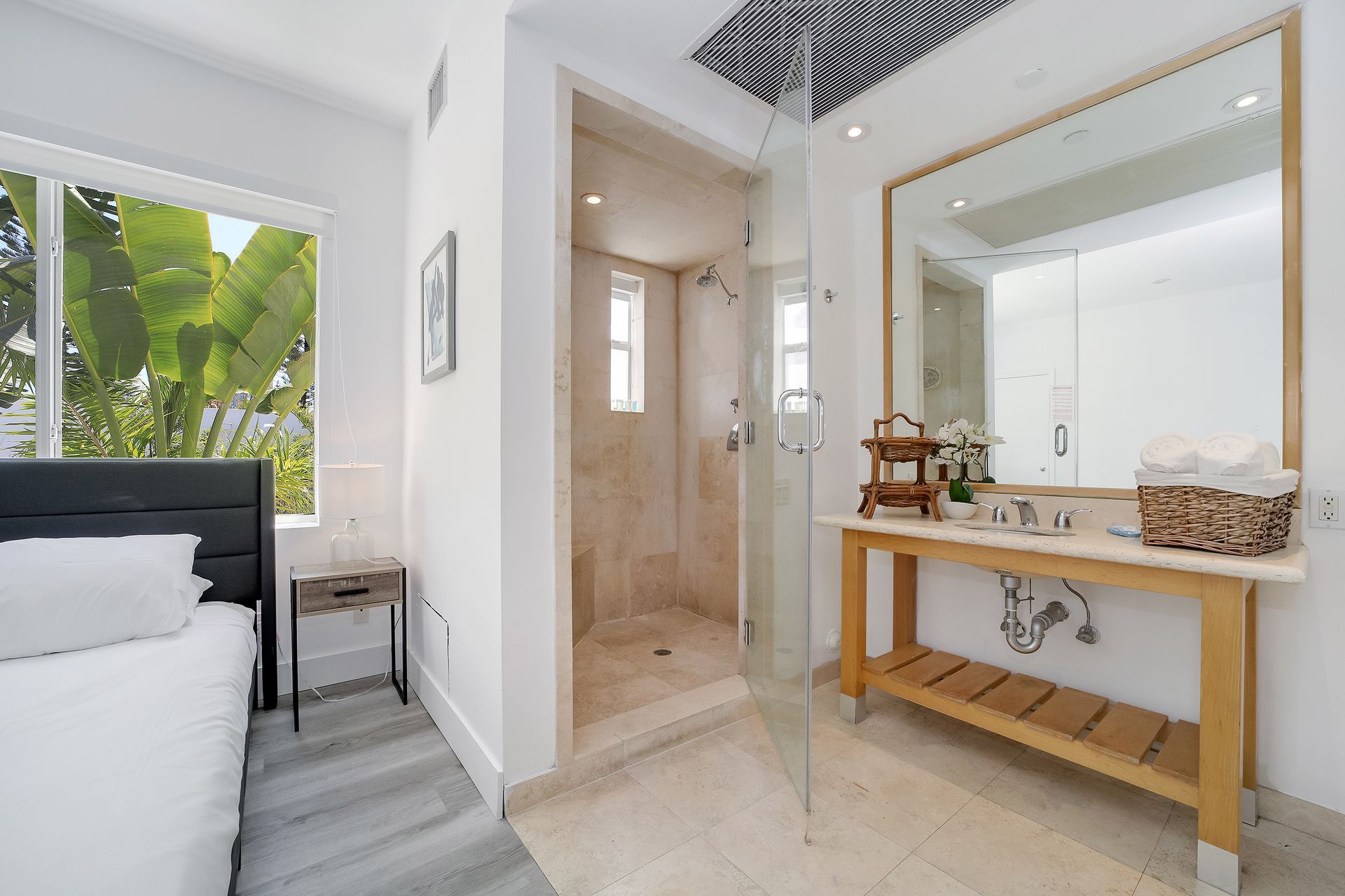 Bedroom with adjacent bathroom: bed by window, shower, vanity with mirror, wood accents.