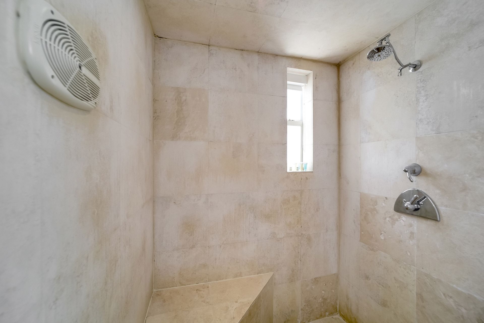 Shower stall with tan stone tiles, built-in bench, small window, and shower fixtures.