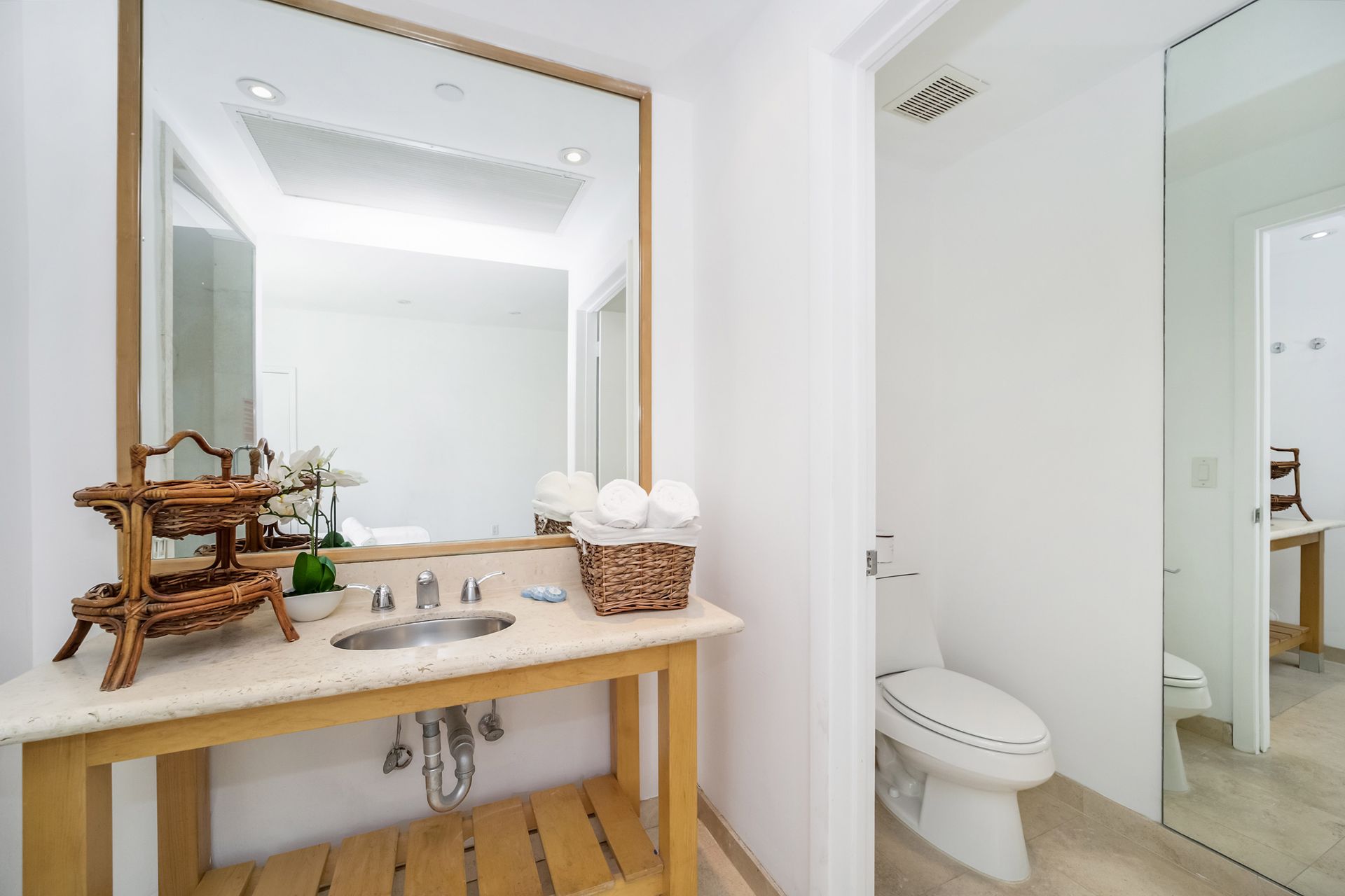 Bathroom with a sink, large mirror, and open doorway to a toilet area; white walls and light-colored counter.
