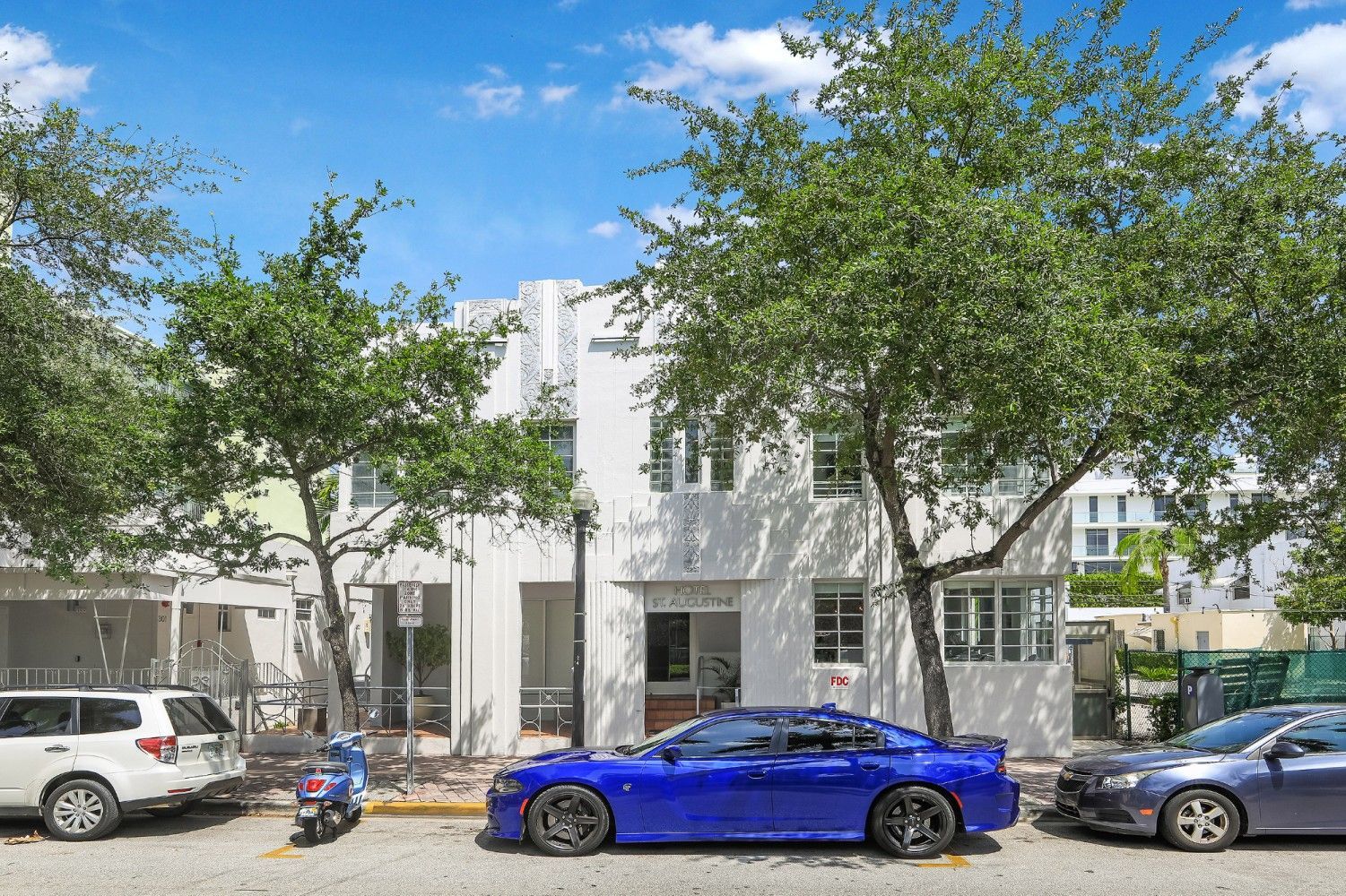 White multi-story building with blue car parked in front, flanked by trees and parked cars.