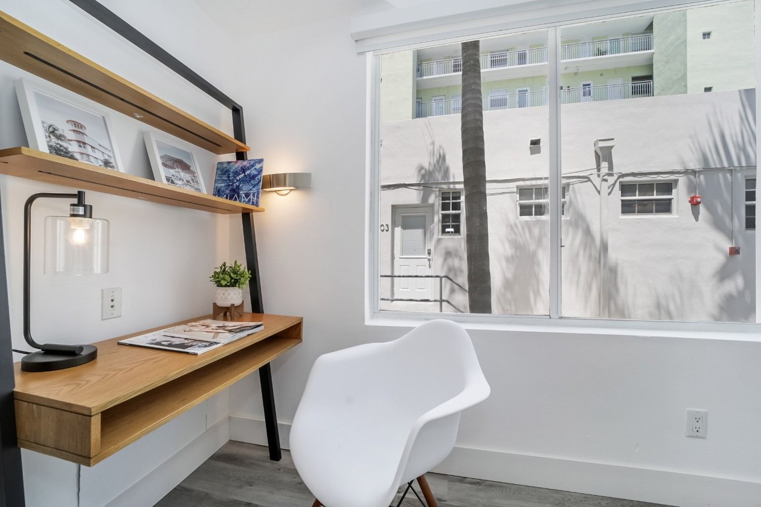Desk with shelves, chair, and window. White walls, wood desk, black lamp.