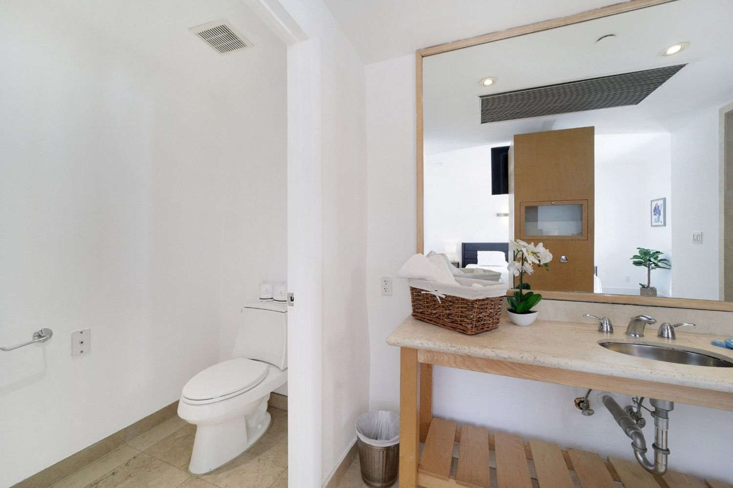 Bathroom with a toilet, sink, and large mirror reflecting another room with a bed and plants.
