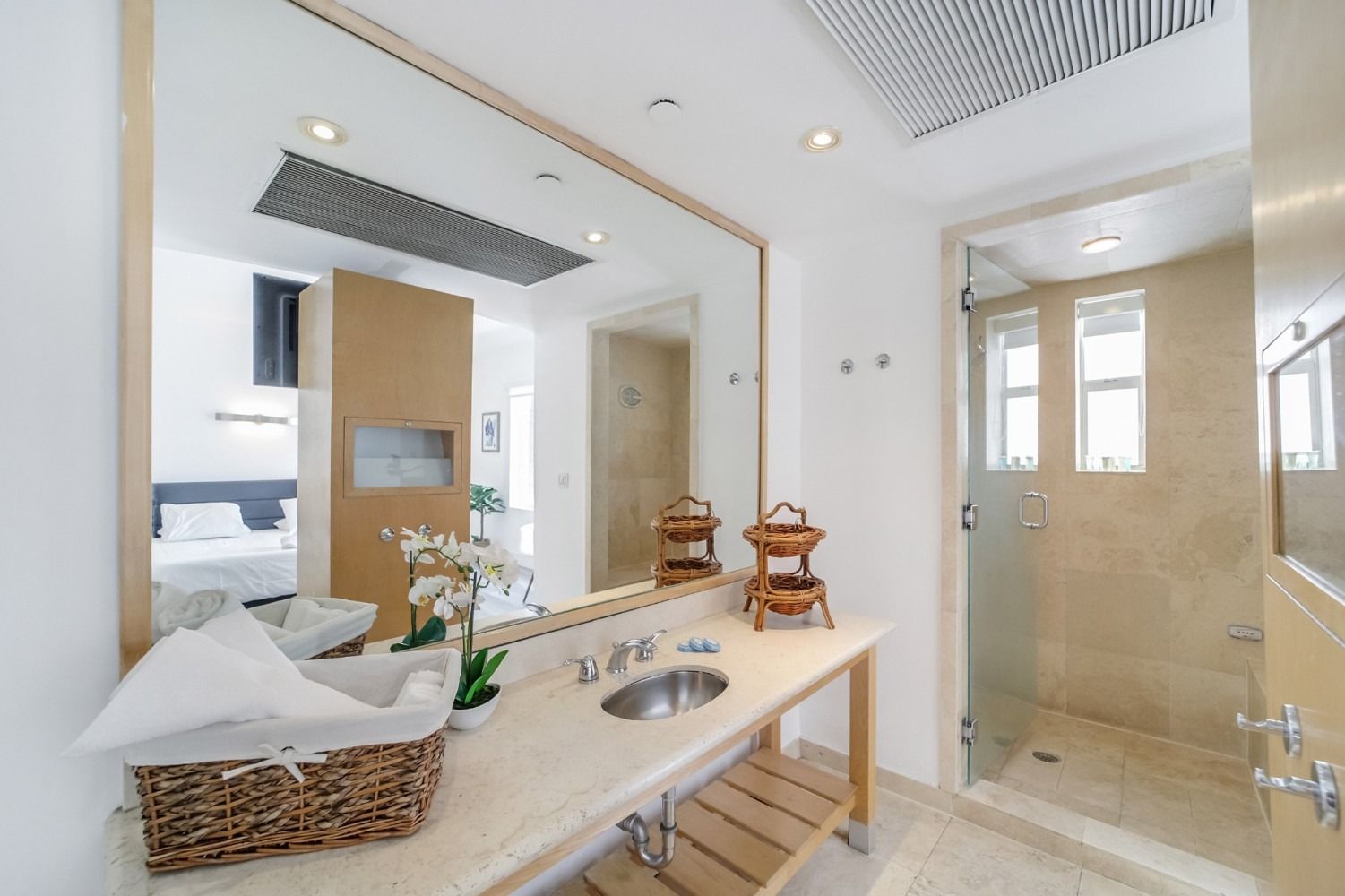 Bathroom with large mirror, sink, and glass shower. White walls, wood accents, and basket with towels.
