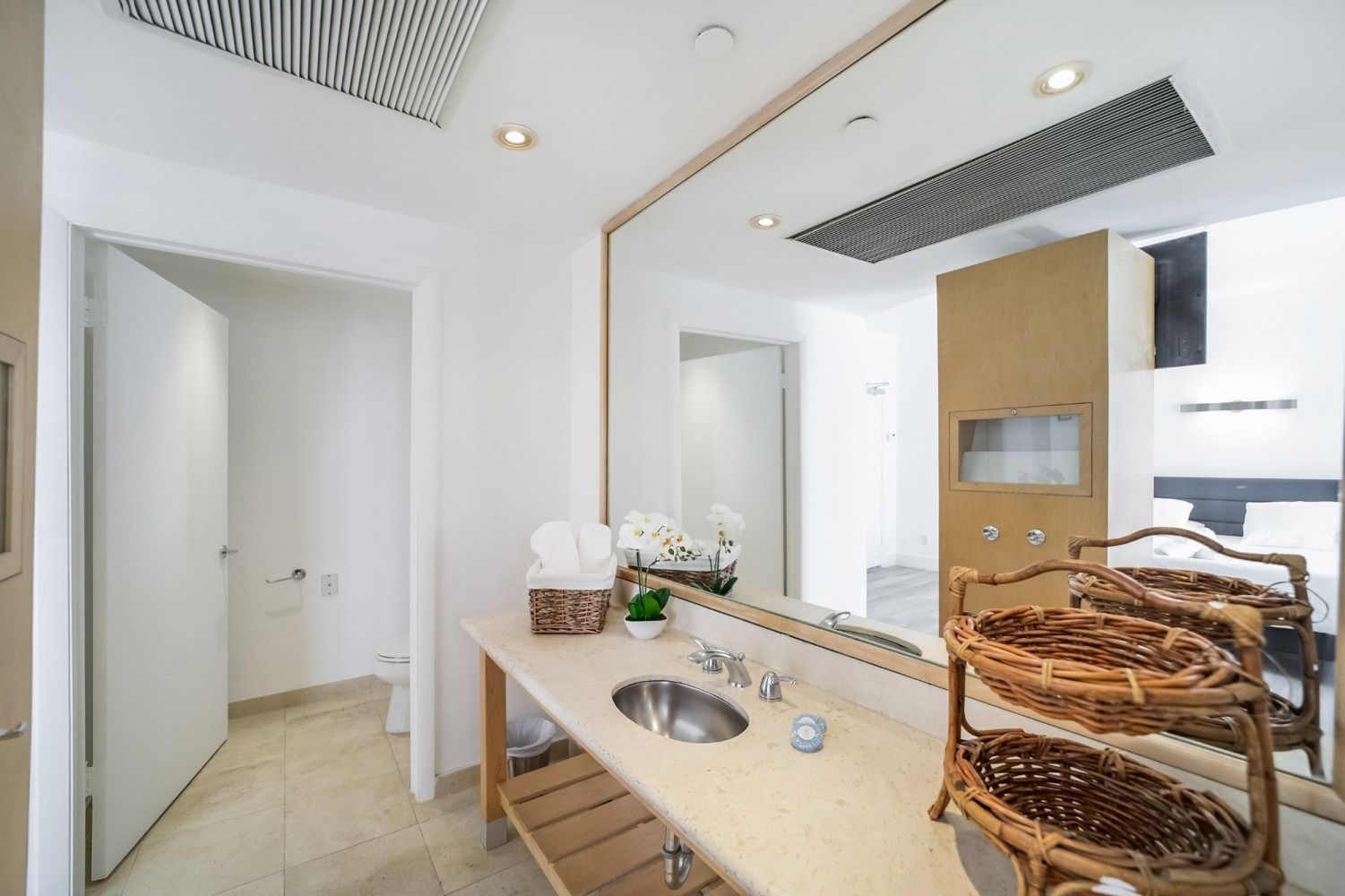 Bathroom with a long mirror, sink, and open door to a toilet area; neutral tones.