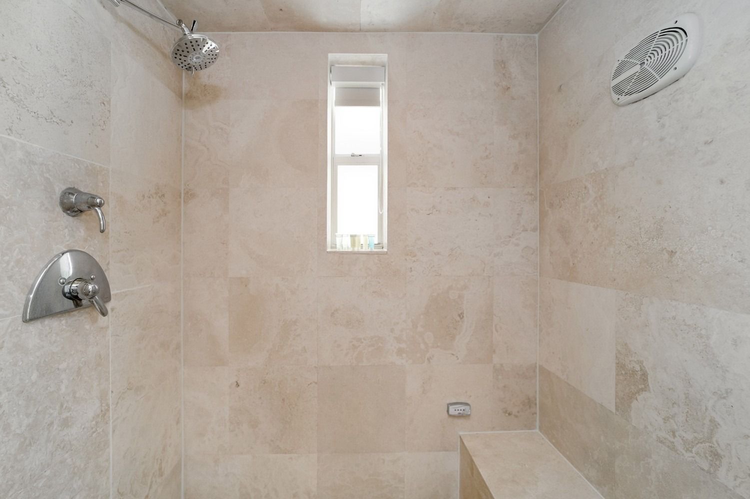 Beige tiled shower with window, shower head, controls, and built-in bench.