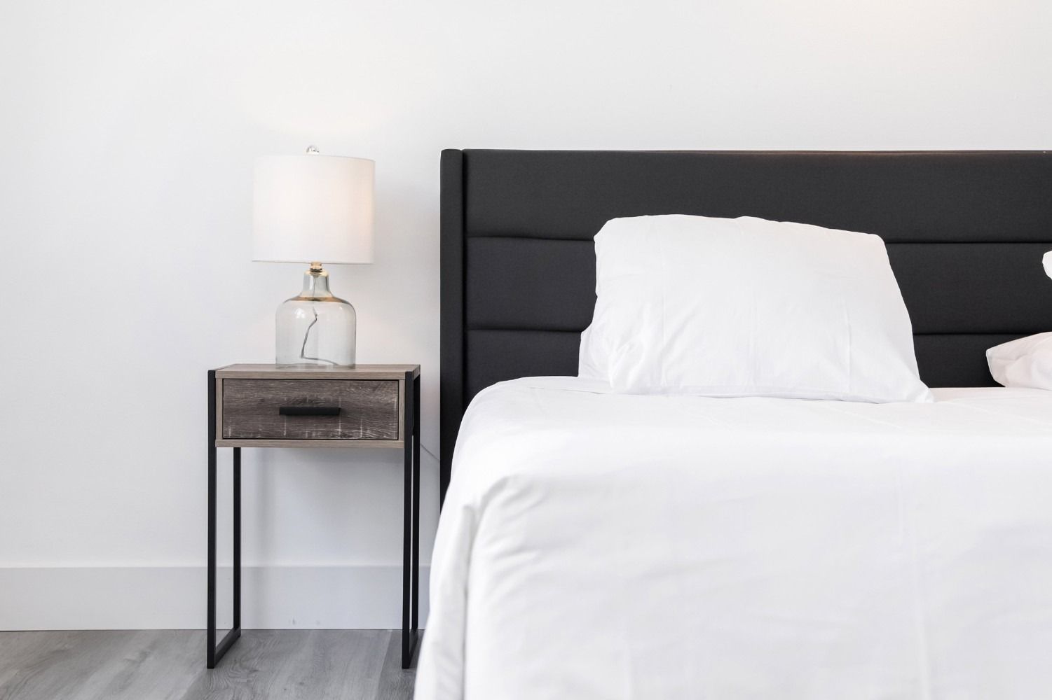 Bed with white bedding and black headboard, next to a nightstand with a lamp.