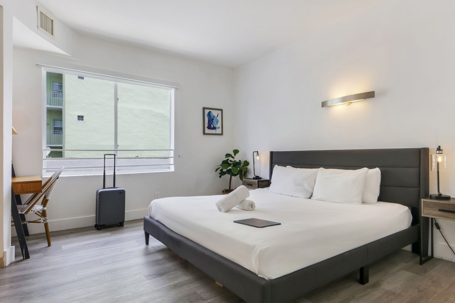 Bedroom with a large bed, suitcase, desk by window, and minimalist decor.