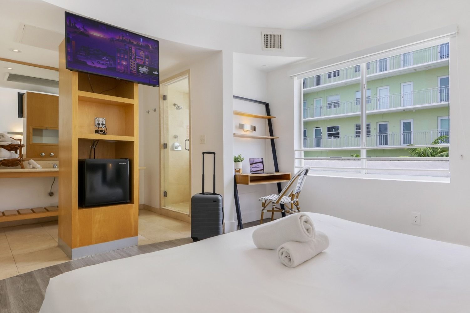 Hotel room with white bed, TV, desk, and bathroom. A suitcase sits by the bed.