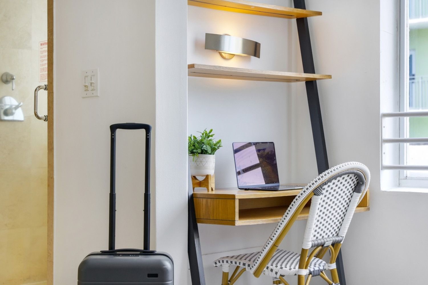 Laptop on desk with suitcase and wicker chair next to window.