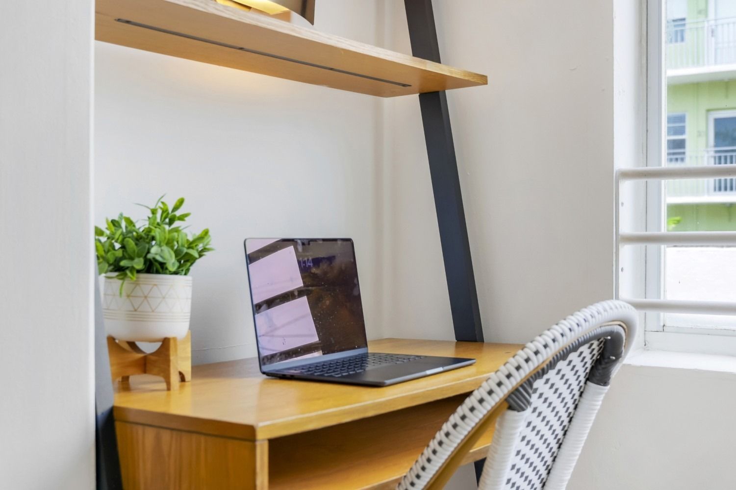 Desk with laptop, plant, and chair by window; wooden shelf above.