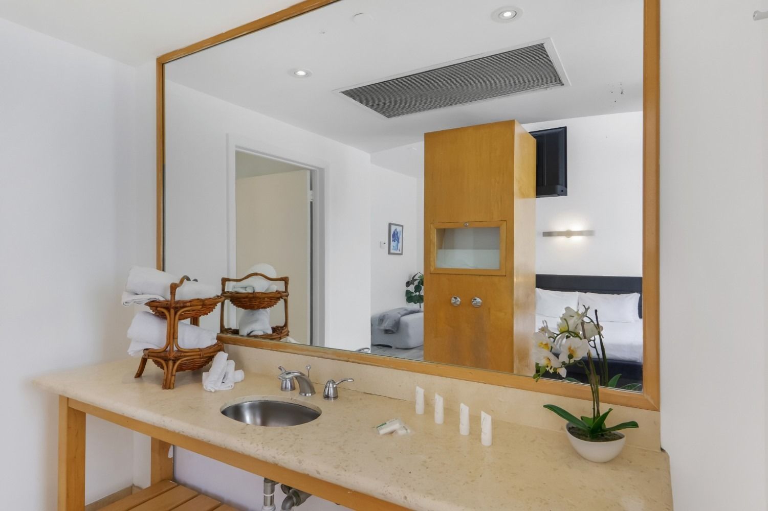 Bathroom with large mirror reflecting a bedroom. Towels, sink, cabinet, and orchids are visible.