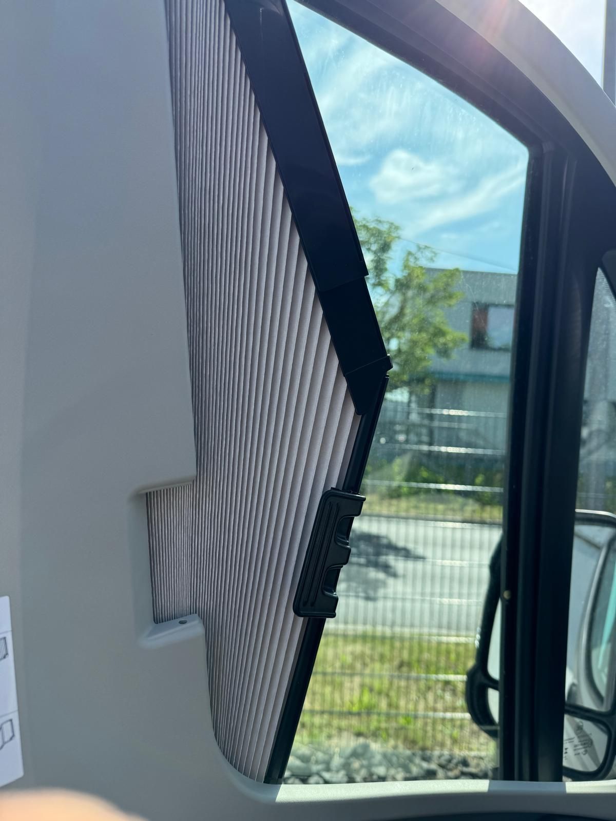 Wohnmobilfenster mit teilweise ausgefahrenem grauen Plissee; sonniger Ausblick nach draußen.