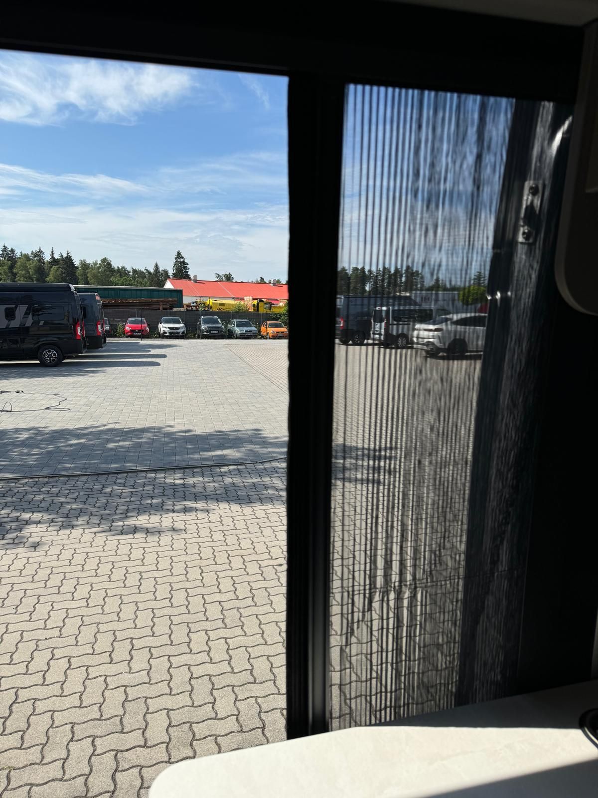 Blick aus dem Fahrzeugfenster auf einen Parkplatz mit Autos und ein Gebäude unter blauem Himmel.